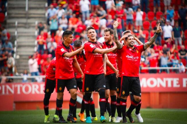 Soccer players in red jerseys celebrating on the field with fans in the background.