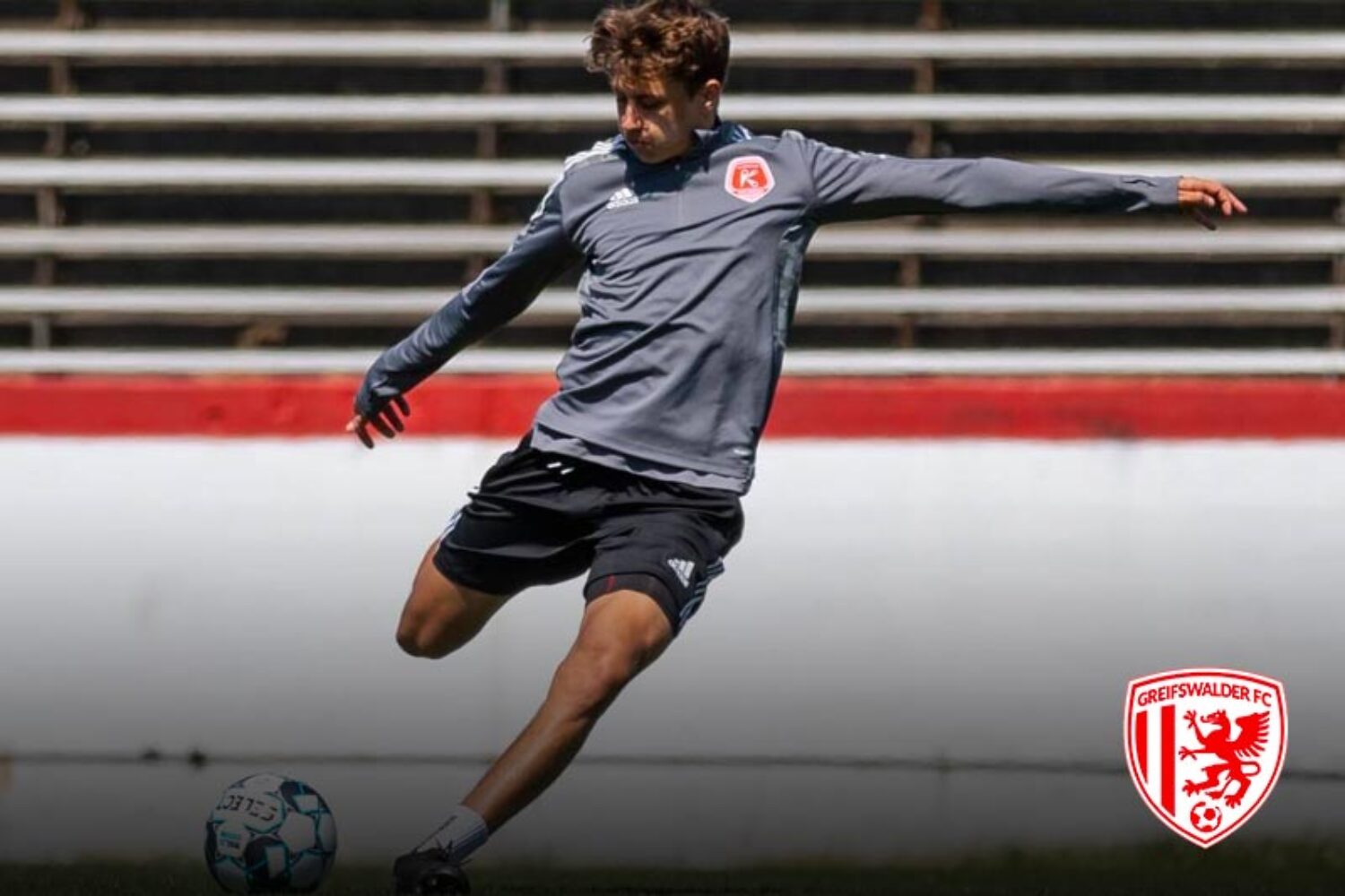 Soccer player in gray jersey kicking ball on field with empty stands.