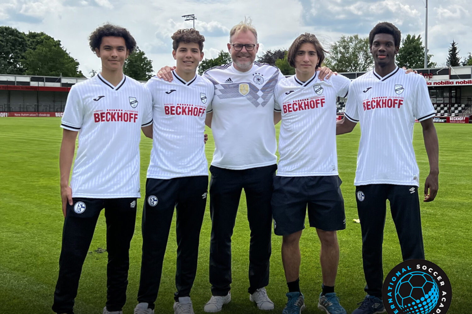 Five people posing on a soccer field, four in matching white jerseys and one in a white sweatshirt.