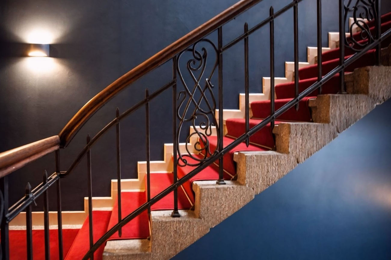 Elegant staircase with red carpet and ornate railing in dim lighting.