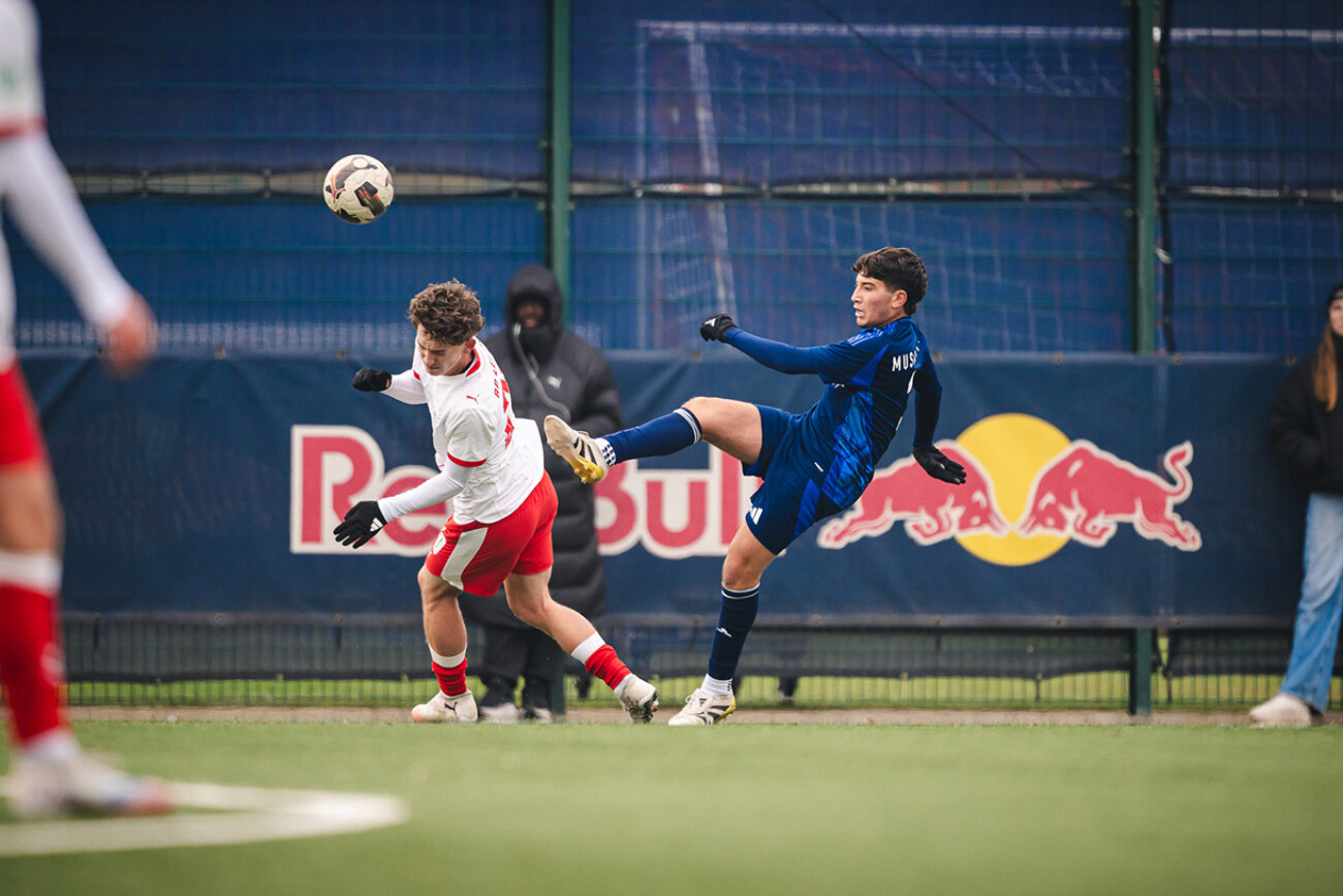 Two young soccer players competing for the ball on a field with a Red Bull banner.