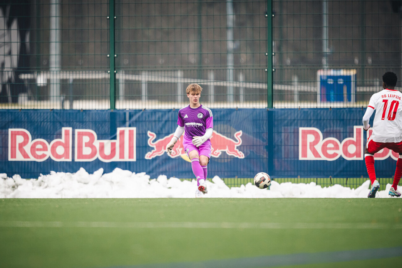 Soccer goalkeeper in purple prepares to kick the ball on a snowy field with Red Bull banners.